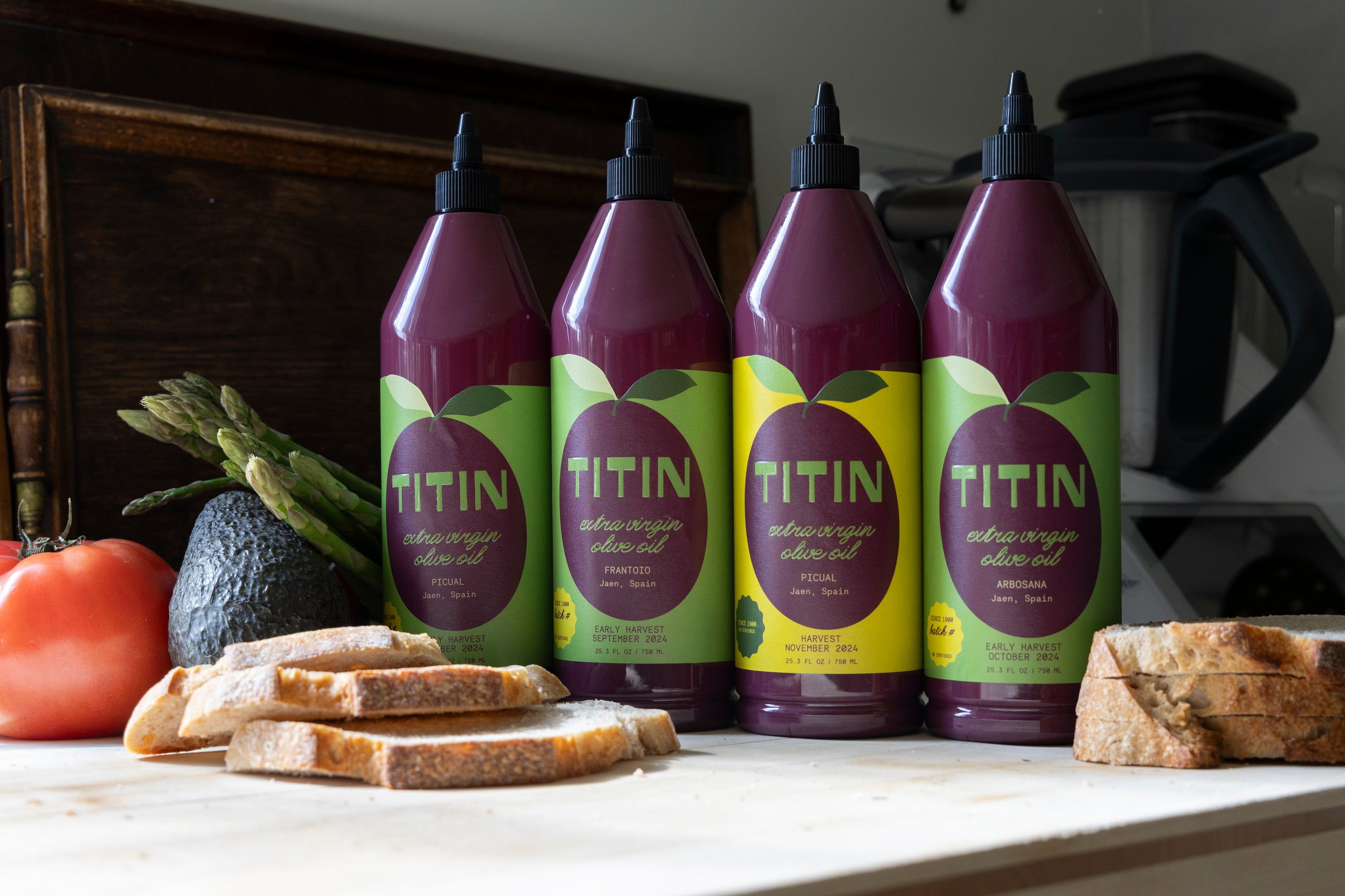 Four bottles of TITIN olive oil on a kitchen counter with bread and vegetables.